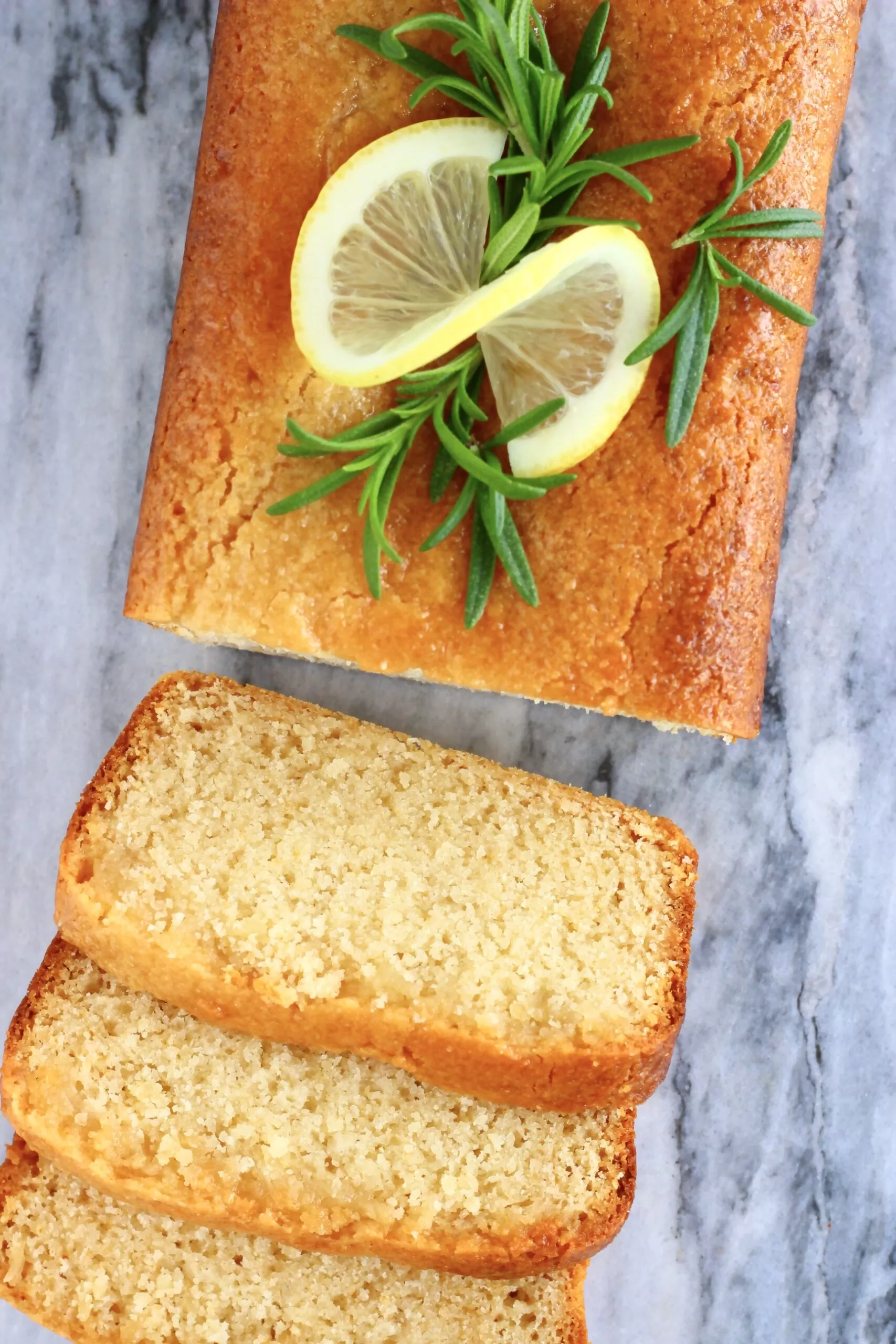 A vegan lemon drizzle loaf cake with three slices next to it on a marble background