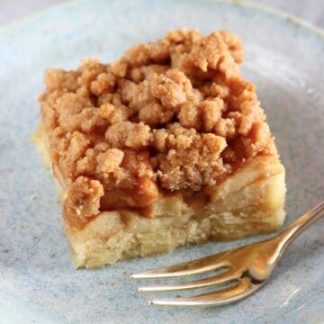 A square piece of vegan apple crumb cake on a plate with a gold fork