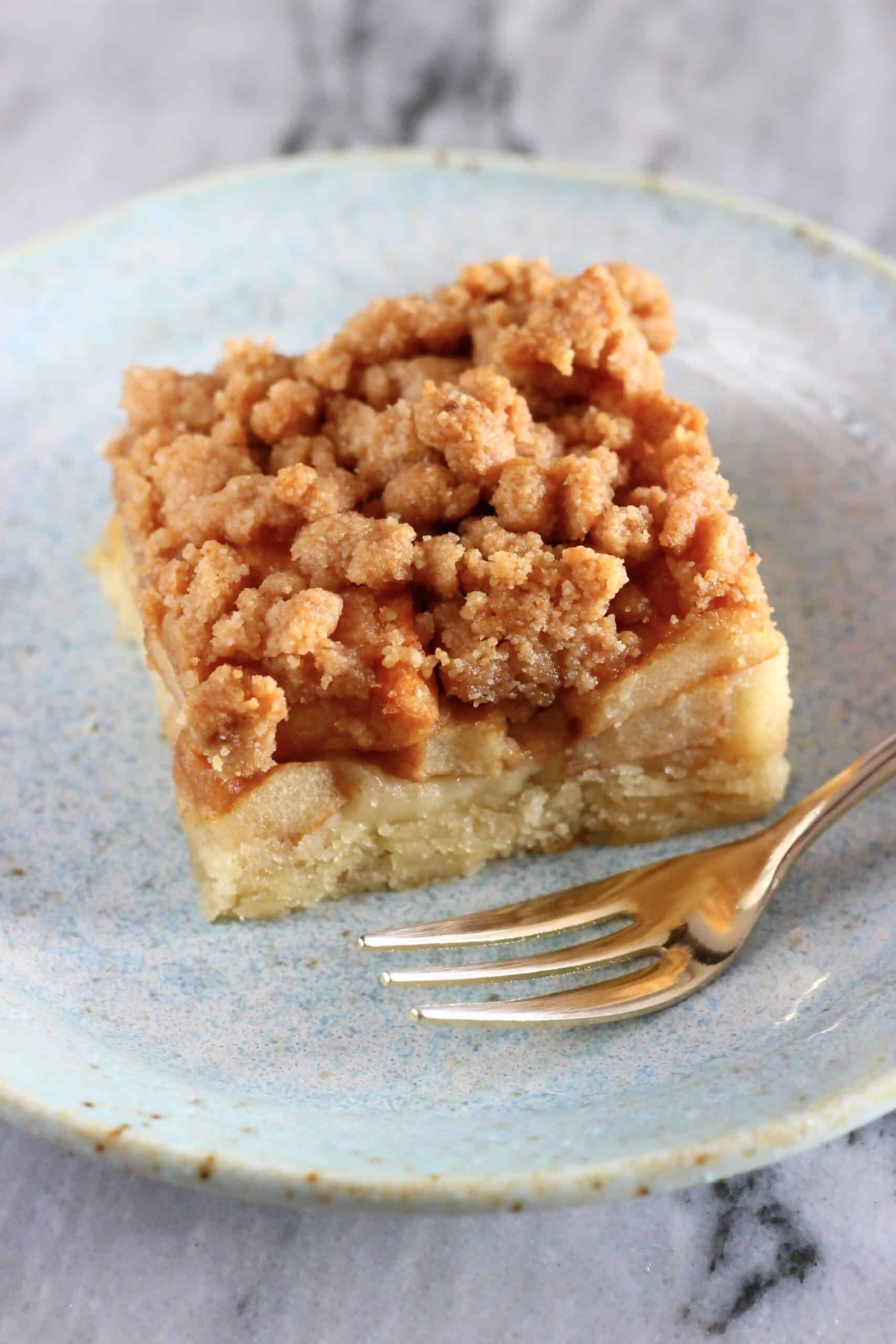 A square piece of vegan apple crumb cake on a plate with a gold fork