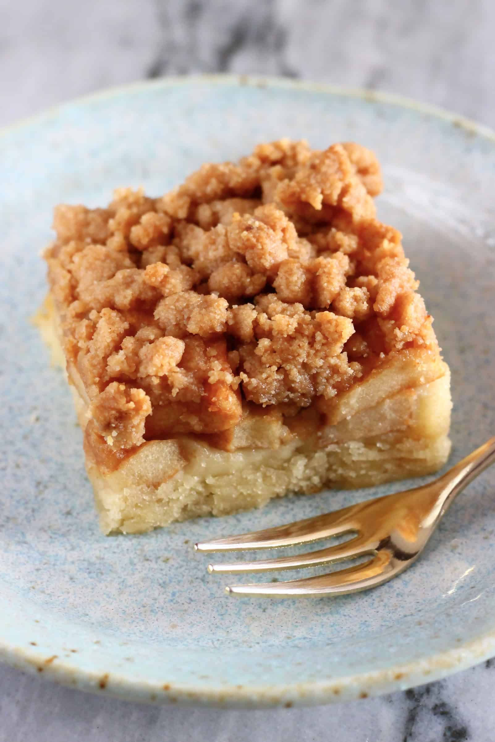 A square piece of vegan apple crumb cake on a plate with a gold fork