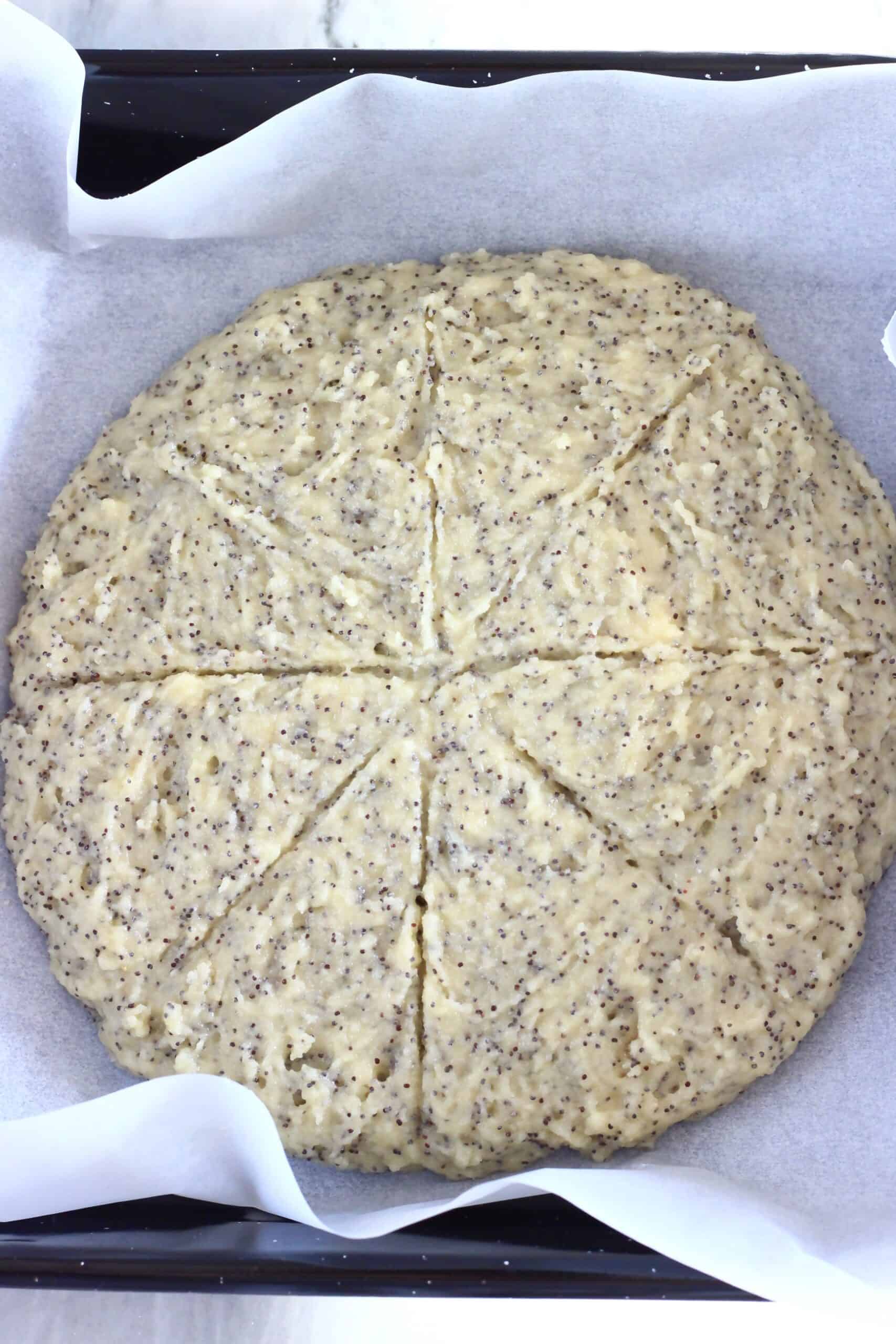 A circle of raw gluten-free vegan lemon poppy seed scones dough scored into eight pieces, on a baking tray lined with baking paper