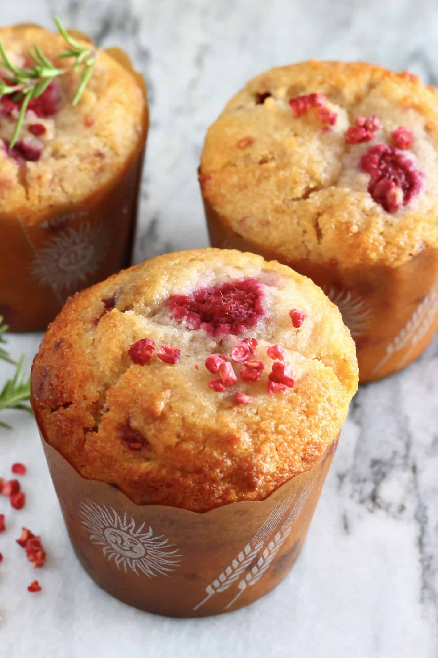 Three gluten-free vegan raspberry muffins in muffin cases on a marble background