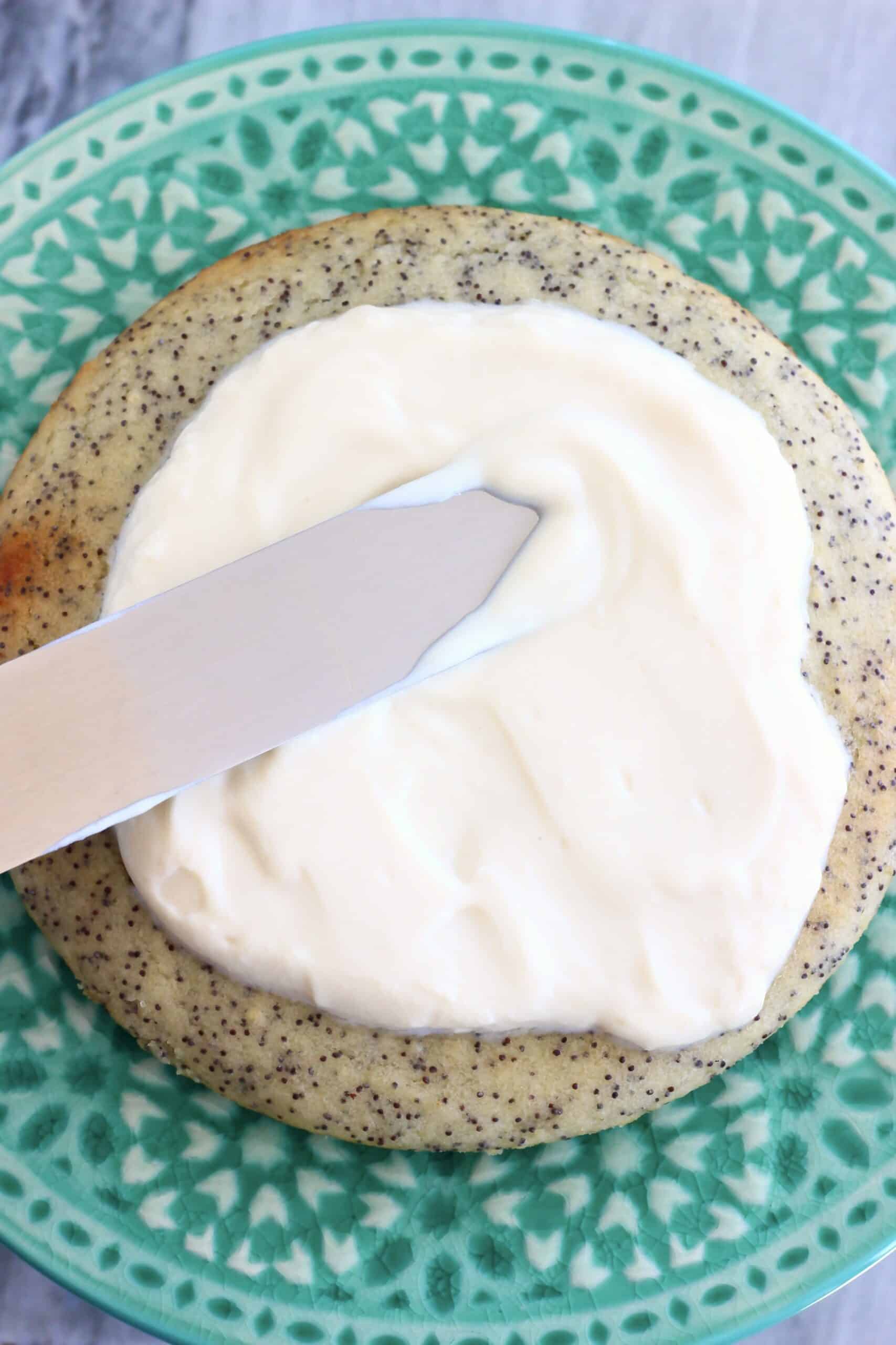 A vegan lemon poppy seed layer cake sponge on a cake stand with a palette knife spreading frosting over the sponge