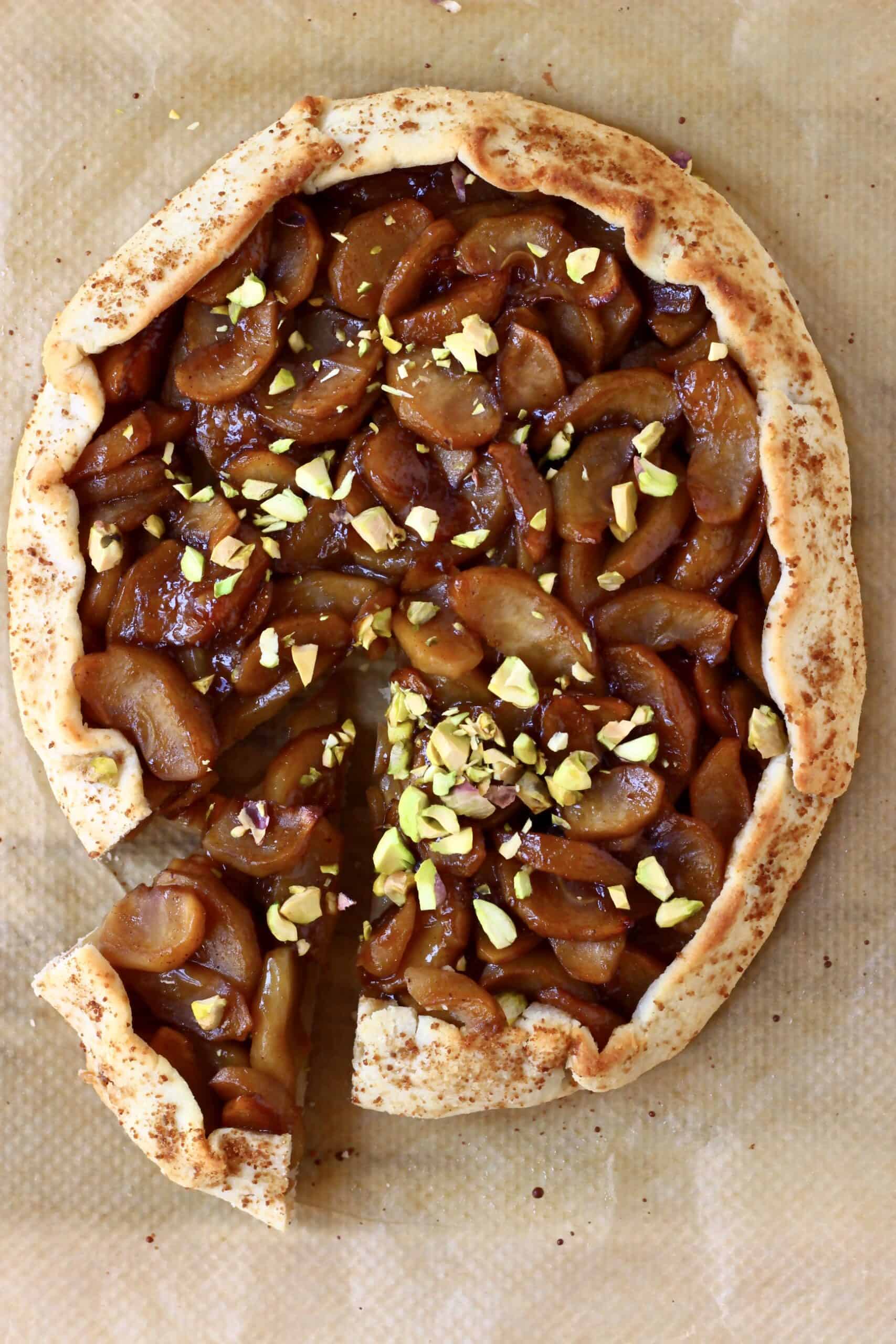 A round galette filled with brown apples sprinkled with pistachio nuts with a slice cut out of it against a sheet of brown baking paper