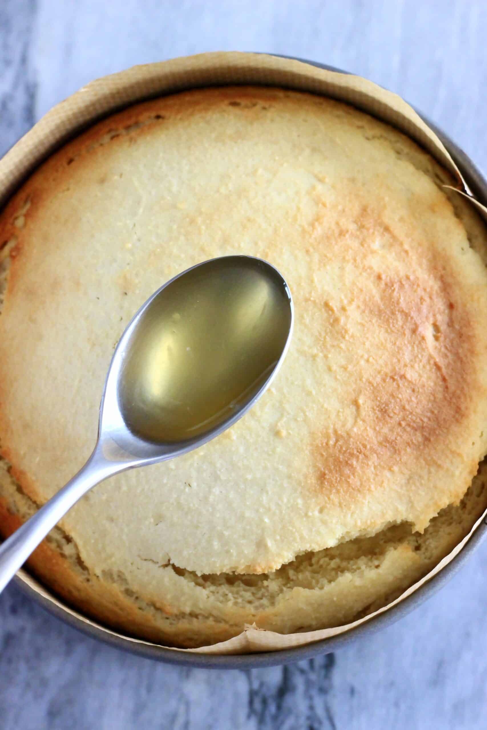 A spoon drizzling lemon syrup over a baked vegan lemon olive oil yogurt cake in a round springform baking tin