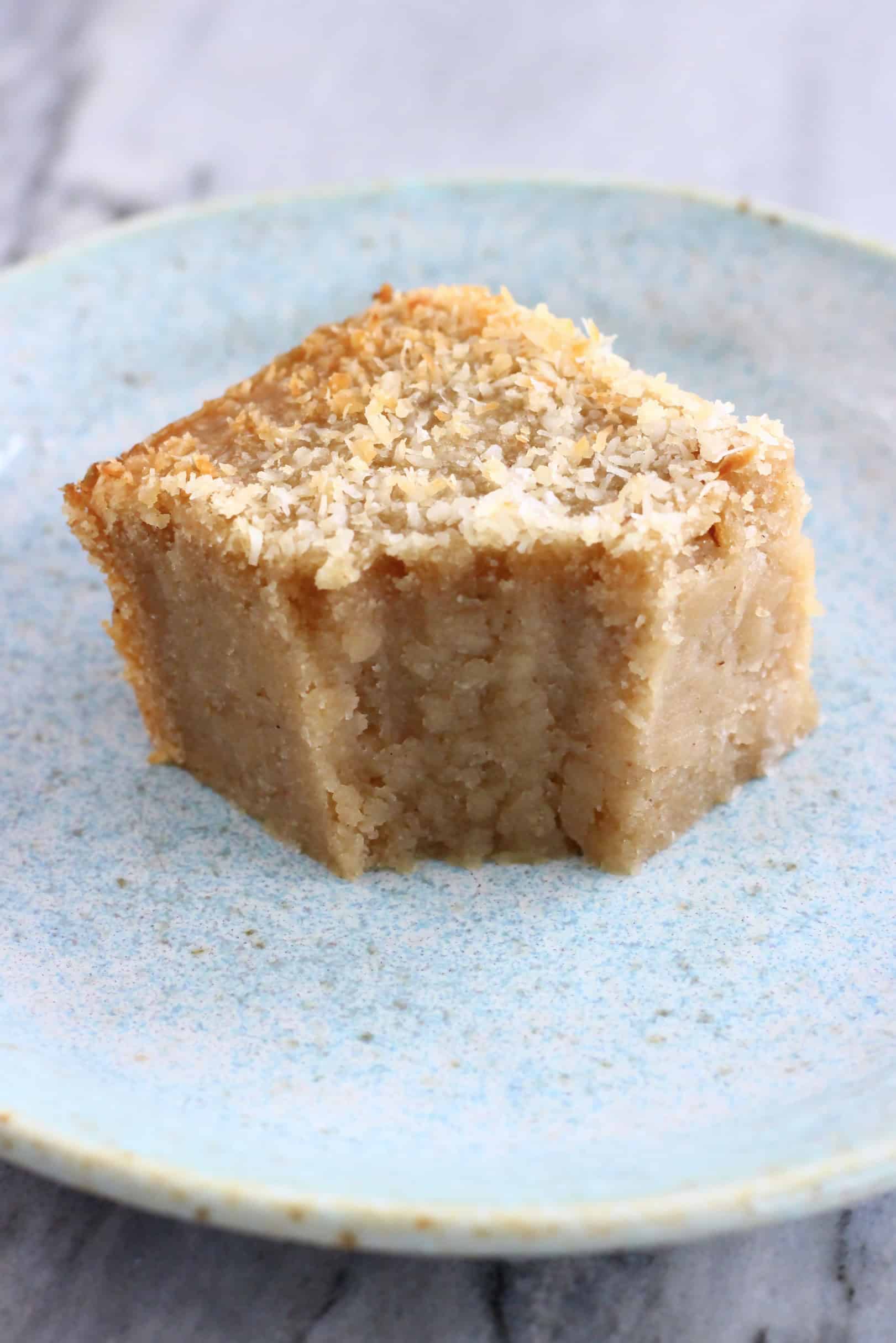 A square of vegan butter mochi topped with desiccated coconut with a bite taken out of it on a plate with a fork