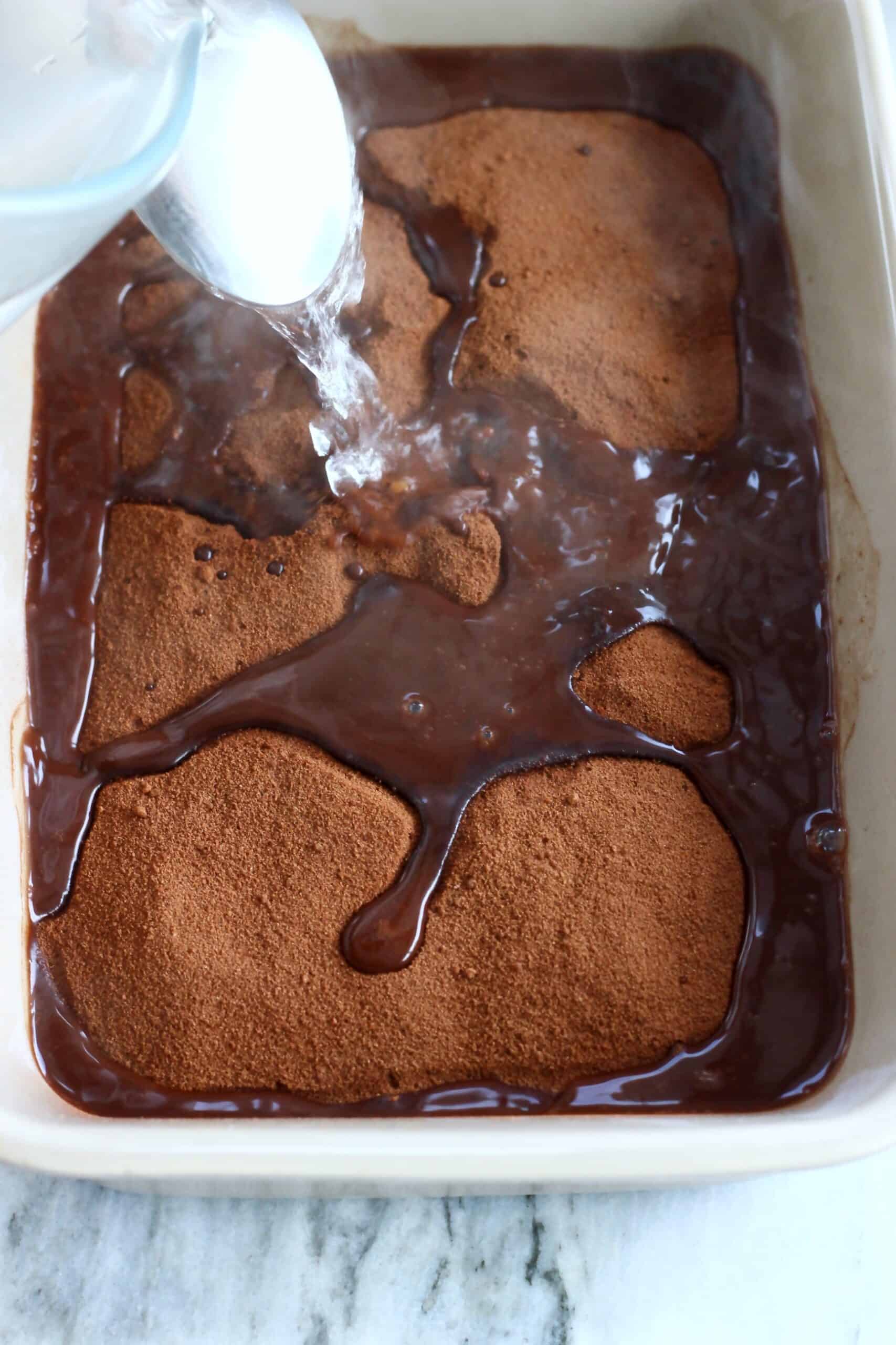 Hot water being poured over vegan chocolate self-saucing pudding batter with cocoa powder and coconut sugar sprinkled on top in a rectangular baking dish
