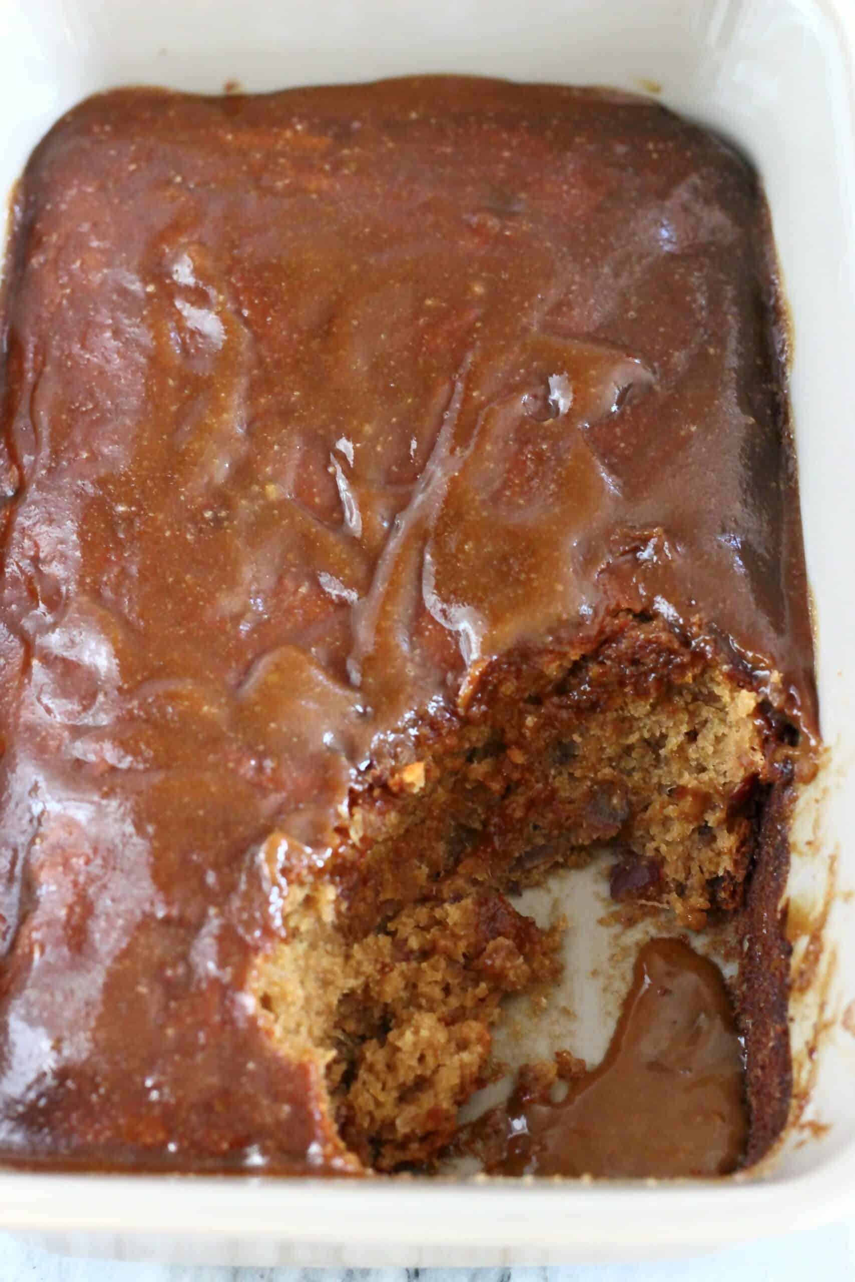 Vegan sticky toffee pudding covered with toffee sauce in a rectangular baking dish with a scoop taken out of it