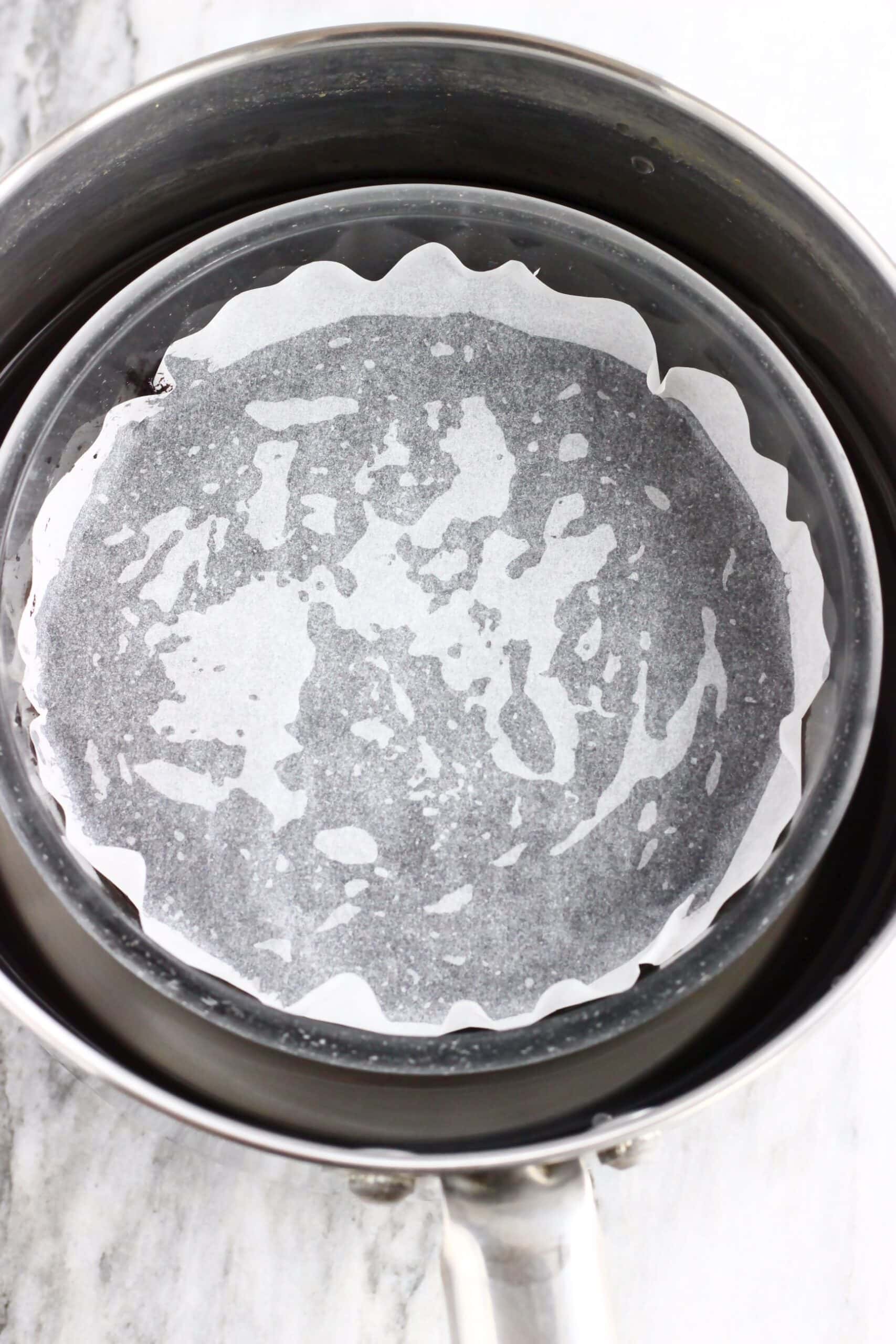 Raw vegan chocolate Christmas pudding batter in a glass bowl in a saucepan with water and a circle of baking paper on top of the batter